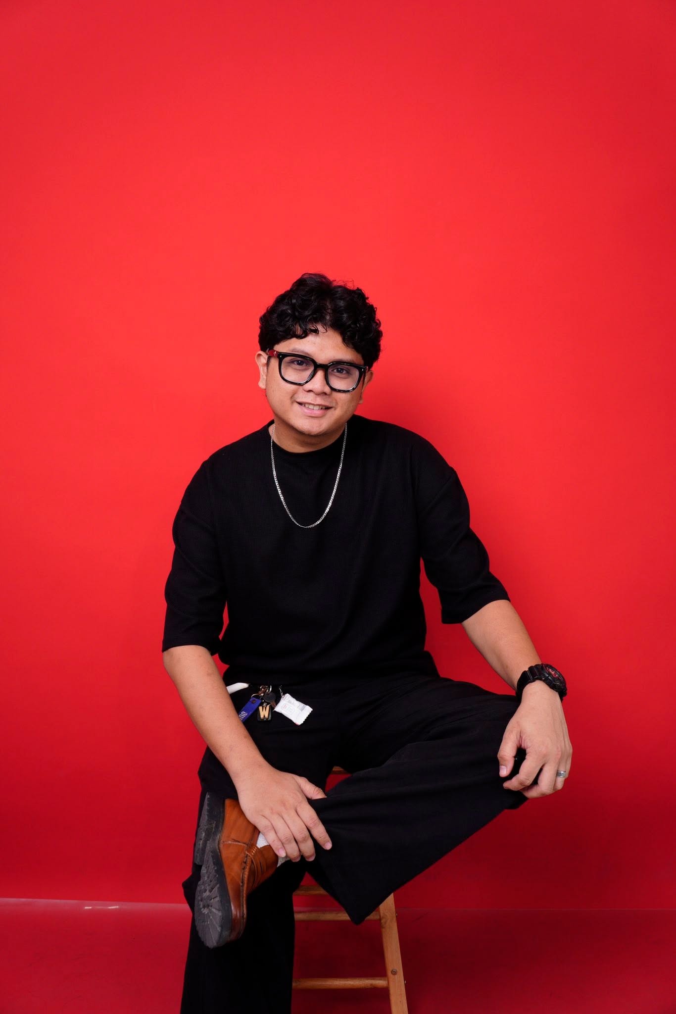 Wacky Jhay Yugto seated on a stool in all black, smiling, against a red backdrop.